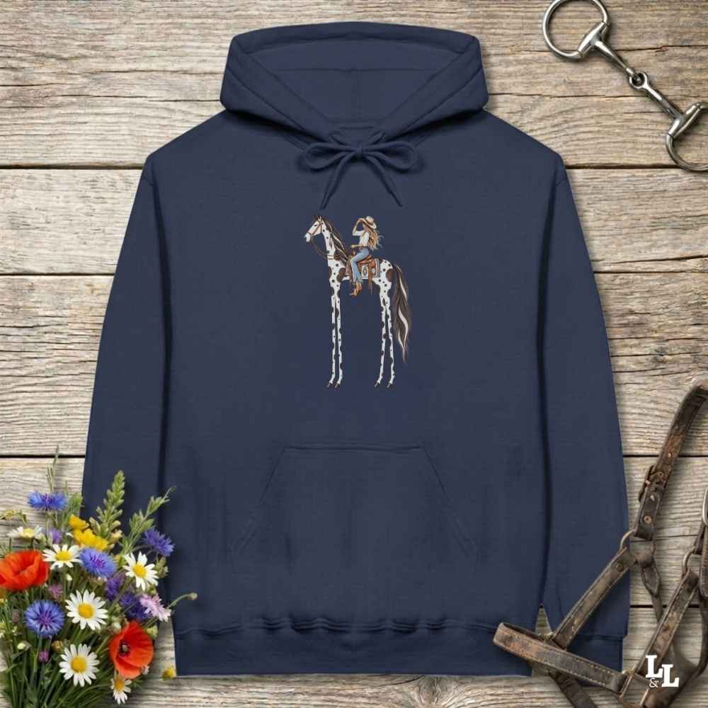 A navy blue pullover hoodie with a graphic of a cowboy riding a horse, displayed on a rustic wooden background with equestrian-themed props like a halter, reins, and a small bouquet of wildflowers.