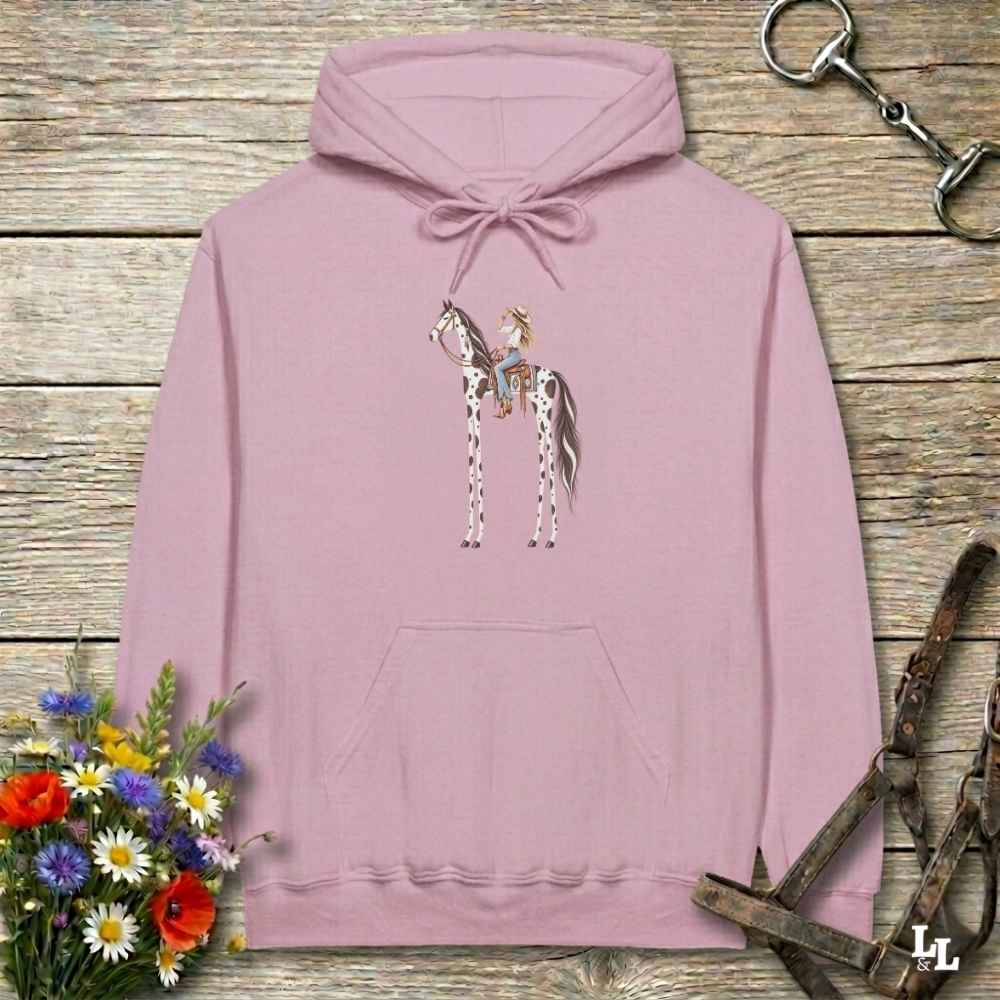 A pink pullover hoodie with a graphic of a cowboy riding a horse, displayed on a rustic wooden background with equestrian-themed props like a halter, reins, and a small bouquet of wildflowers.
