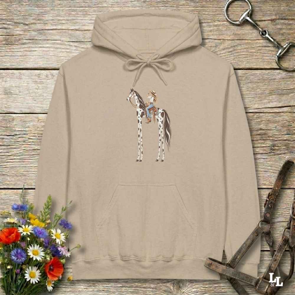 A beige pullover hoodie with a graphic of a cowboy riding a horse, displayed on a rustic wooden background with equestrian-themed props like a halter, reins, and a small bouquet of wildflowers.