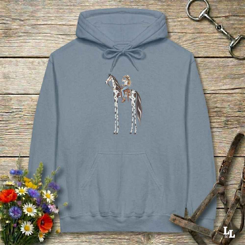 A blue pullover hoodie with a graphic of a cowboy riding a horse, displayed on a rustic wooden background with equestrian-themed props like a halter, reins, and a small bouquet of wildflowers.