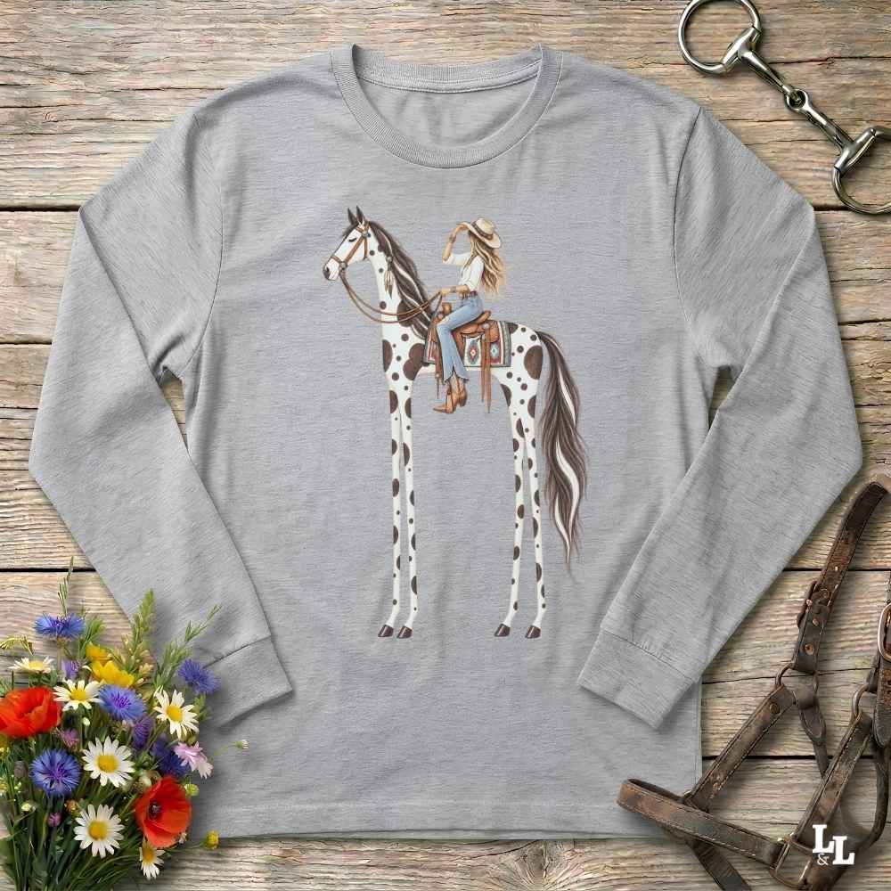A heather gray long-sleeve t-shirt with a graphic of a woman riding a tall horse, styled in a flat-lay on a rustic wooden background with a horse bit, leather bridle, and a small bouquet of wildflowers.