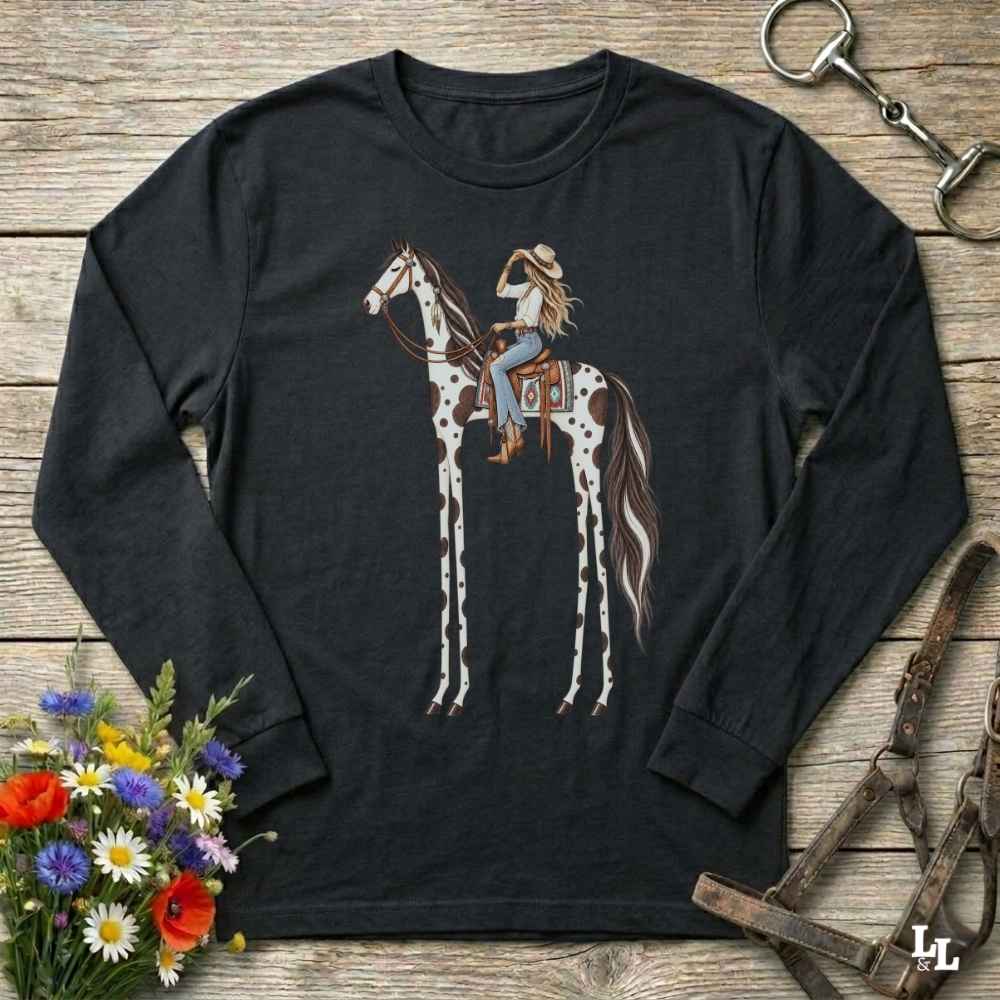 A black long-sleeve t-shirt with a graphic of a woman riding a tall horse, styled in a flat-lay on a rustic wooden background with a horse bit, leather bridle, and a small bouquet of wildflowers.