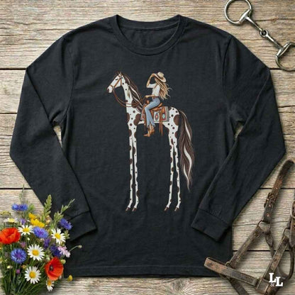 A black long-sleeve t-shirt with a graphic of a woman riding a tall horse, styled in a flat-lay on a rustic wooden background with a horse bit, leather bridle, and a small bouquet of wildflowers.