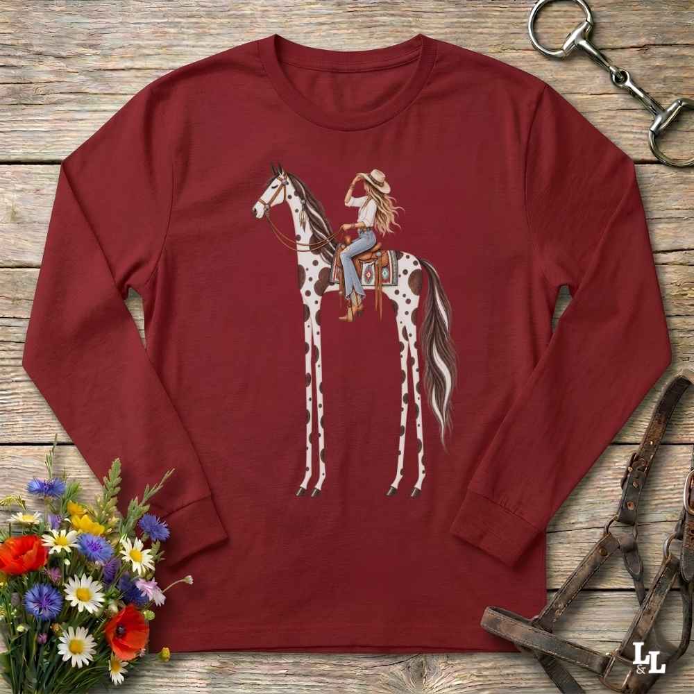 A red long-sleeve t-shirt with a graphic of a woman riding a tall horse, styled in a flat-lay on a rustic wooden background with a horse bit, leather bridle, and a small bouquet of wildflowers.