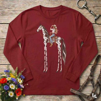 A red long-sleeve t-shirt with a graphic of a woman riding a tall horse, styled in a flat-lay on a rustic wooden background with a horse bit, leather bridle, and a small bouquet of wildflowers.