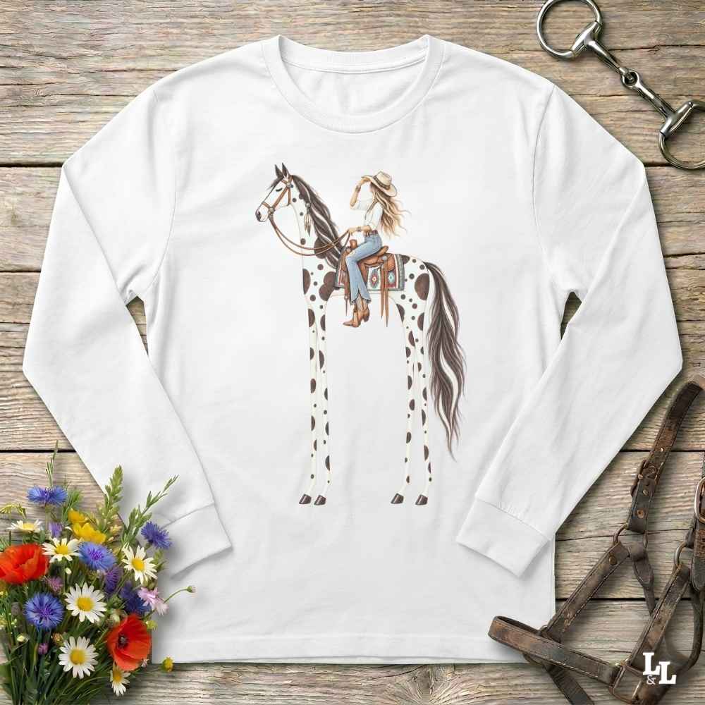A white  long-sleeve t-shirt with a graphic of a woman riding a tall horse, styled in a flat-lay on a rustic wooden background with a horse bit, leather bridle, and a small bouquet of wildflowers.