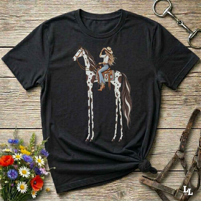 A black short-sleeve graphic T-shirt laid flat on a rustic wooden surface, featuring an illustration of a woman riding a tall spotted horse. The shirt is styled with flowers, a horse bit, and a leather bridle around it.
