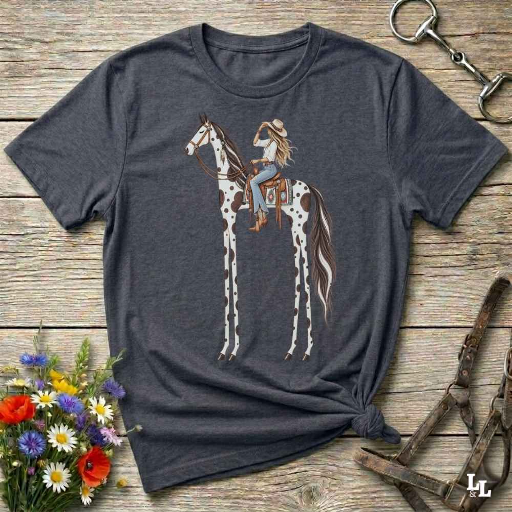 A grey short-sleeve graphic T-shirt laid flat on a rustic wooden surface, featuring an illustration of a woman riding a tall spotted horse. The shirt is styled with flowers, a horse bit, and a leather bridle around it.
