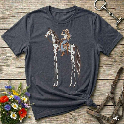 A grey short-sleeve graphic T-shirt laid flat on a rustic wooden surface, featuring an illustration of a woman riding a tall spotted horse. The shirt is styled with flowers, a horse bit, and a leather bridle around it.