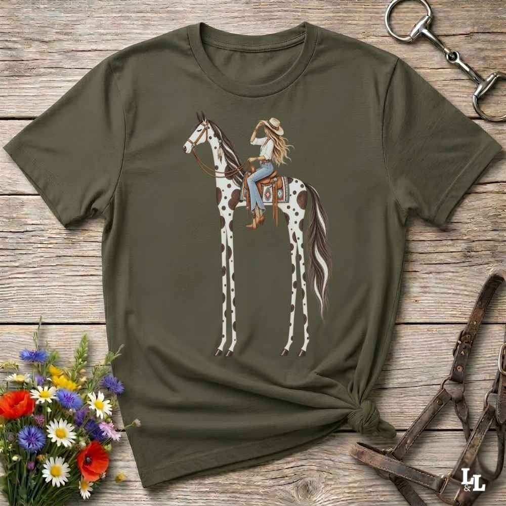 A green short-sleeve graphic T-shirt laid flat on a rustic wooden surface, featuring an illustration of a woman riding a tall spotted horse. The shirt is styled with flowers, a horse bit, and a leather bridle around it.