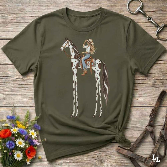 A green short-sleeve graphic T-shirt laid flat on a rustic wooden surface, featuring an illustration of a woman riding a tall spotted horse. The shirt is styled with flowers, a horse bit, and a leather bridle around it.