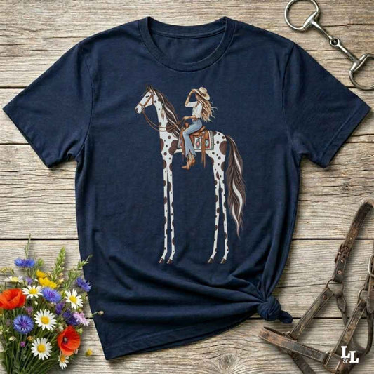 A navy blue short-sleeve t-shirt with a graphic of a cowboy riding a spotted giraffe horse, styled in a flat-lay on a rustic wooden background with flowers and horse tack props.