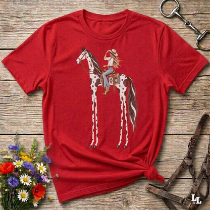 A red short-sleeve graphic T-shirt laid flat on a rustic wooden surface, featuring an illustration of a woman riding a tall spotted horse. The shirt is styled with flowers, a horse bit, and a leather bridle around it.