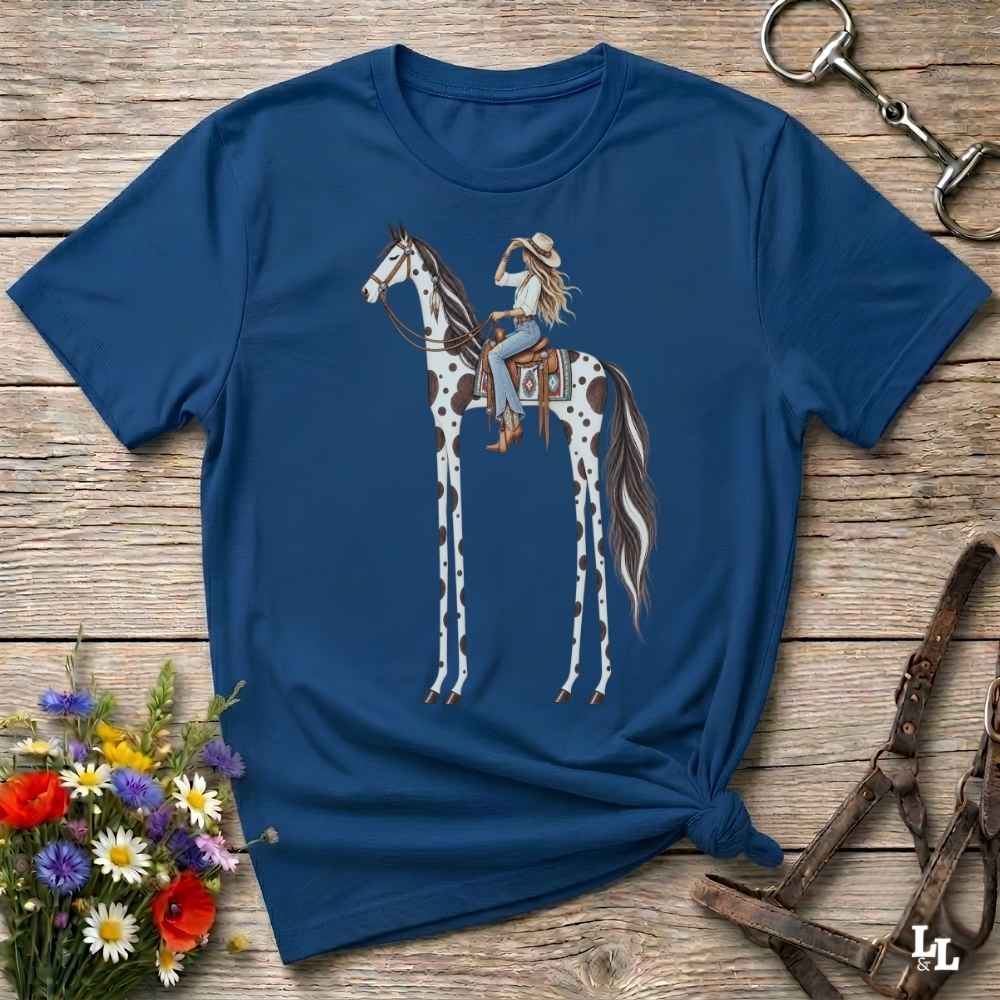 A blue short-sleeve graphic T-shirt laid flat on a rustic wooden surface, featuring an illustration of a woman riding a tall spotted horse. The shirt is styled with flowers, a horse bit, and a leather bridle around it.