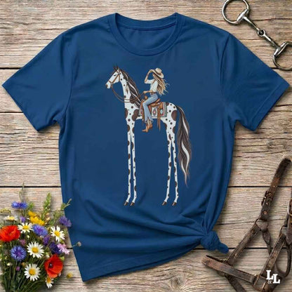 A blue short-sleeve graphic T-shirt laid flat on a rustic wooden surface, featuring an illustration of a woman riding a tall spotted horse. The shirt is styled with flowers, a horse bit, and a leather bridle around it.
