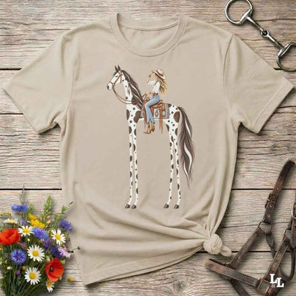 A beige short-sleeve graphic T-shirt laid flat on a rustic wooden surface, featuring an illustration of a woman riding a tall spotted horse. The shirt is styled with flowers, a horse bit, and a leather bridle around it.