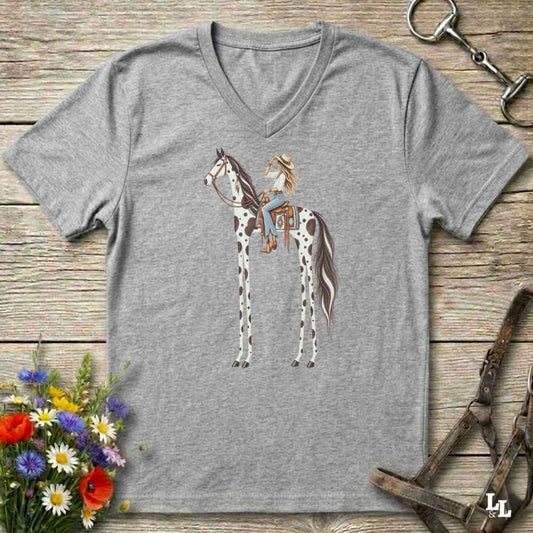A heather gray V-neck t-shirt featuring a graphic of a woman riding a horse with long legs, styled in a flat-lay on a rustic wooden background with equestrian accessories and flowers.