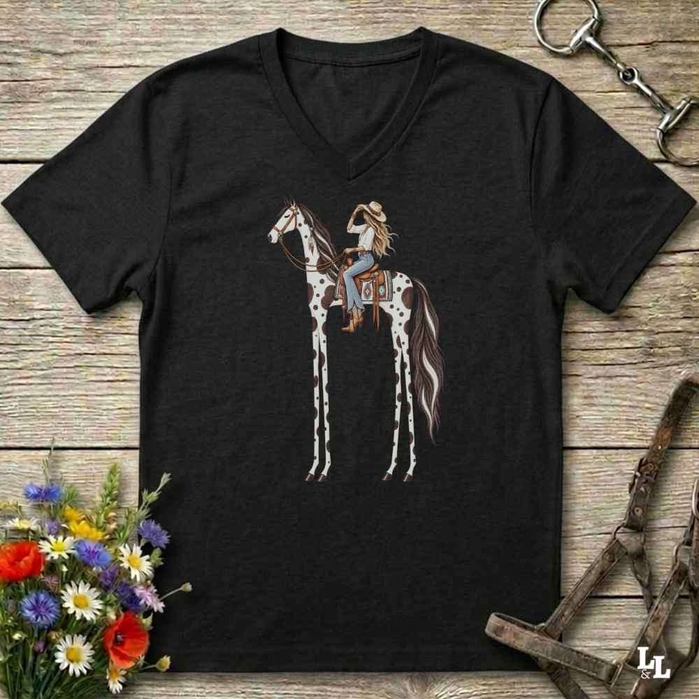 A black V-neck t-shirt laid flat on a rustic wooden surface, featuring a graphic of a woman riding a tall, spotted horse. The shirt is styled with equestrian accessories (a halter and bit) and a small bouquet of wildflowers.
