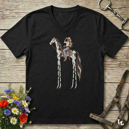 A black V-neck t-shirt laid flat on a rustic wooden surface, featuring a graphic of a woman riding a tall, spotted horse. The shirt is styled with equestrian accessories (a halter and bit) and a small bouquet of wildflowers.