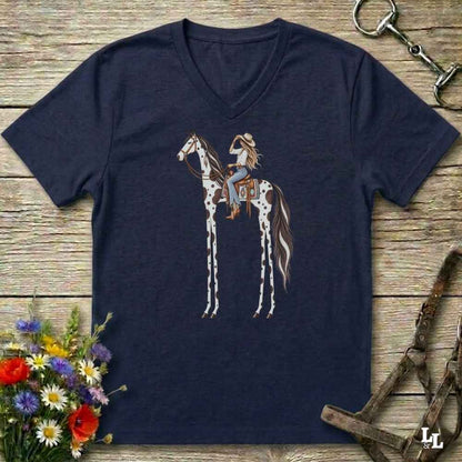 A navy blue V-neck t-shirt laid flat on a rustic wooden surface, featuring a graphic of a woman riding a tall, thin-spotted horse. The shirt is styled with equestrian accessories (a halter and bit) and a small bouquet of wildflowers.