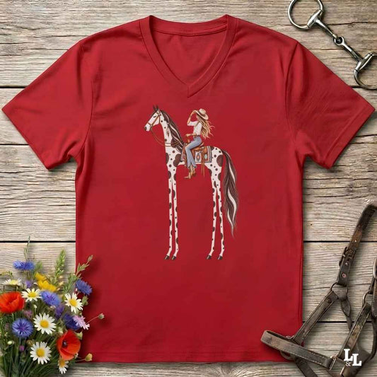 A red V-neck t-shirt laid flat on a rustic wooden surface, featuring a graphic of a woman riding a tall, spotted horse. The shirt is styled with equestrian accessories (a halter and bit) and a small bouquet of wildflowers.