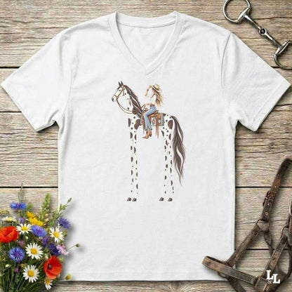 A white V-neck t-shirt laid flat on a rustic wooden surface, featuring a graphic of a woman riding a tall, spotted horse. The shirt is styled with equestrian accessories (a halter and bit) and a small bouquet of wildflowers.