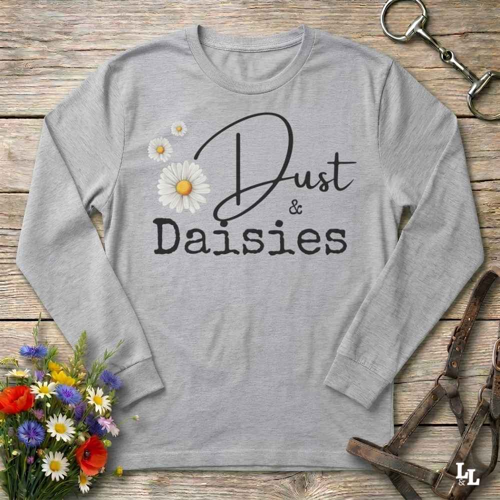 A light heather gray long-sleeve t-shirt with a graphic print reading 'Dust & Daisies' in black script and serif fonts, decorated with three white daisy flowers with yellow centers. The shirt is laid flat on a rustic wooden background with decorative equestrian tack and a small flower bouquet.