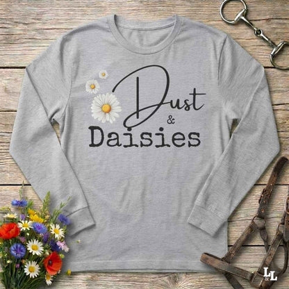 A light heather gray long-sleeve t-shirt with a graphic print reading 'Dust & Daisies' in black script and serif fonts, decorated with three white daisy flowers with yellow centers. The shirt is laid flat on a rustic wooden background with decorative equestrian tack and a small flower bouquet.