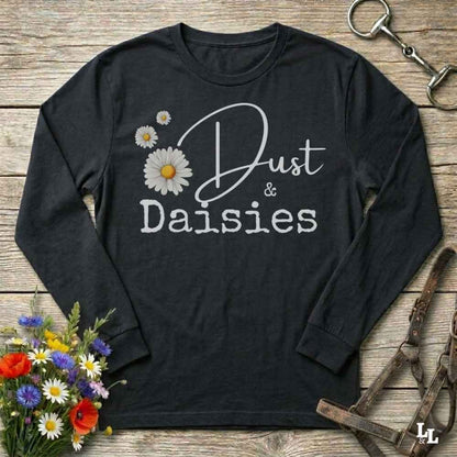 A black long-sleeve t-shirt with a graphic print reading 'Dust & Daisies' in white script and block lettering, decorated with three white daisy flowers with yellow centers. The shirt is displayed in a flat-lay arrangement on a rustic wooden background, surrounded by equestrian-themed accessories (a horse bit, leather bridle, and a bouquet of colorful wildflowers).