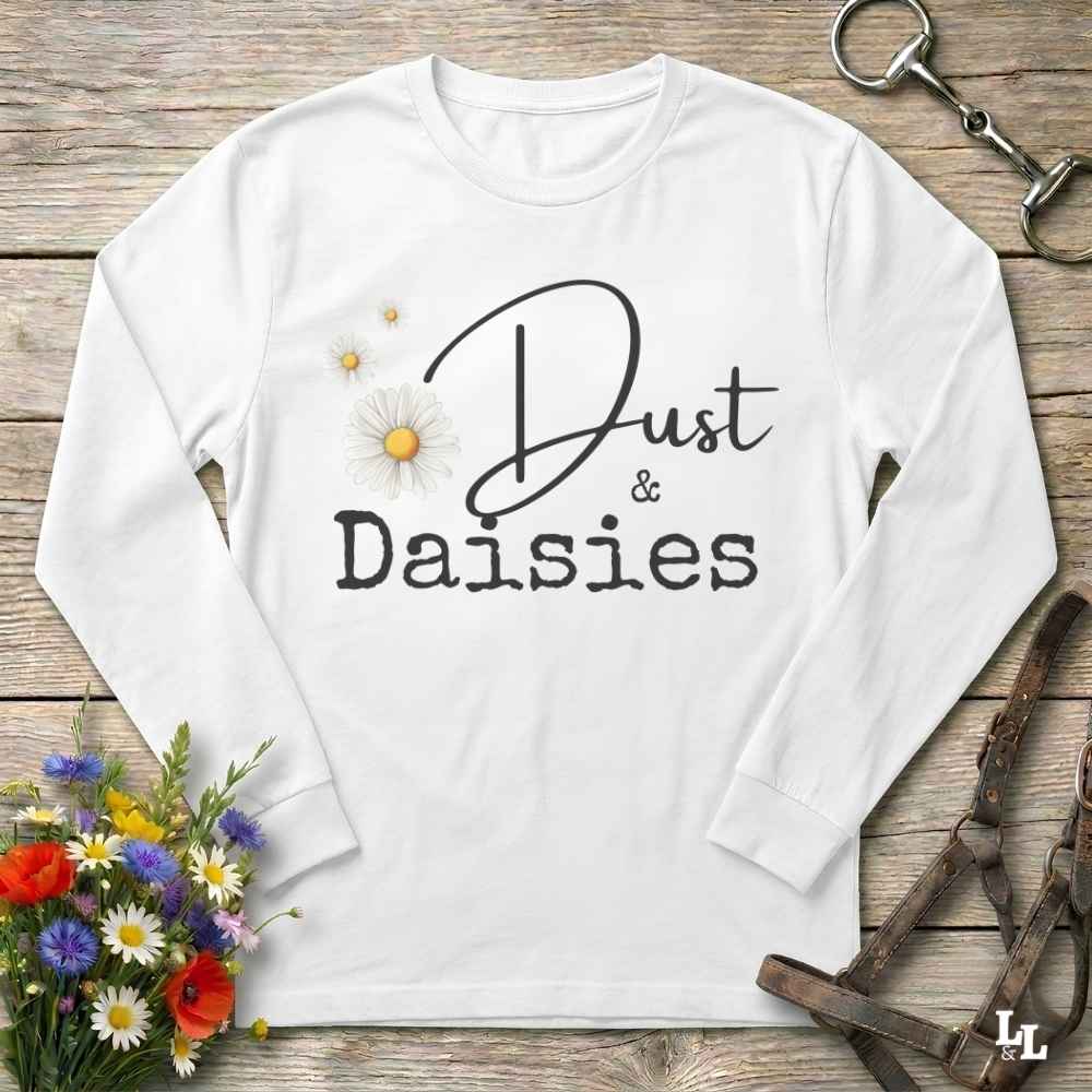 A white long-sleeve t-shirt with a graphic print reading 'Dust & Daisies' in black script and serif fonts, decorated with a daisy illustration. The shirt is displayed in a flat-lay arrangement on a rustic wooden background, surrounded by equestrian-themed accessories (bridle, reins) and a small bouquet of wildflowers.