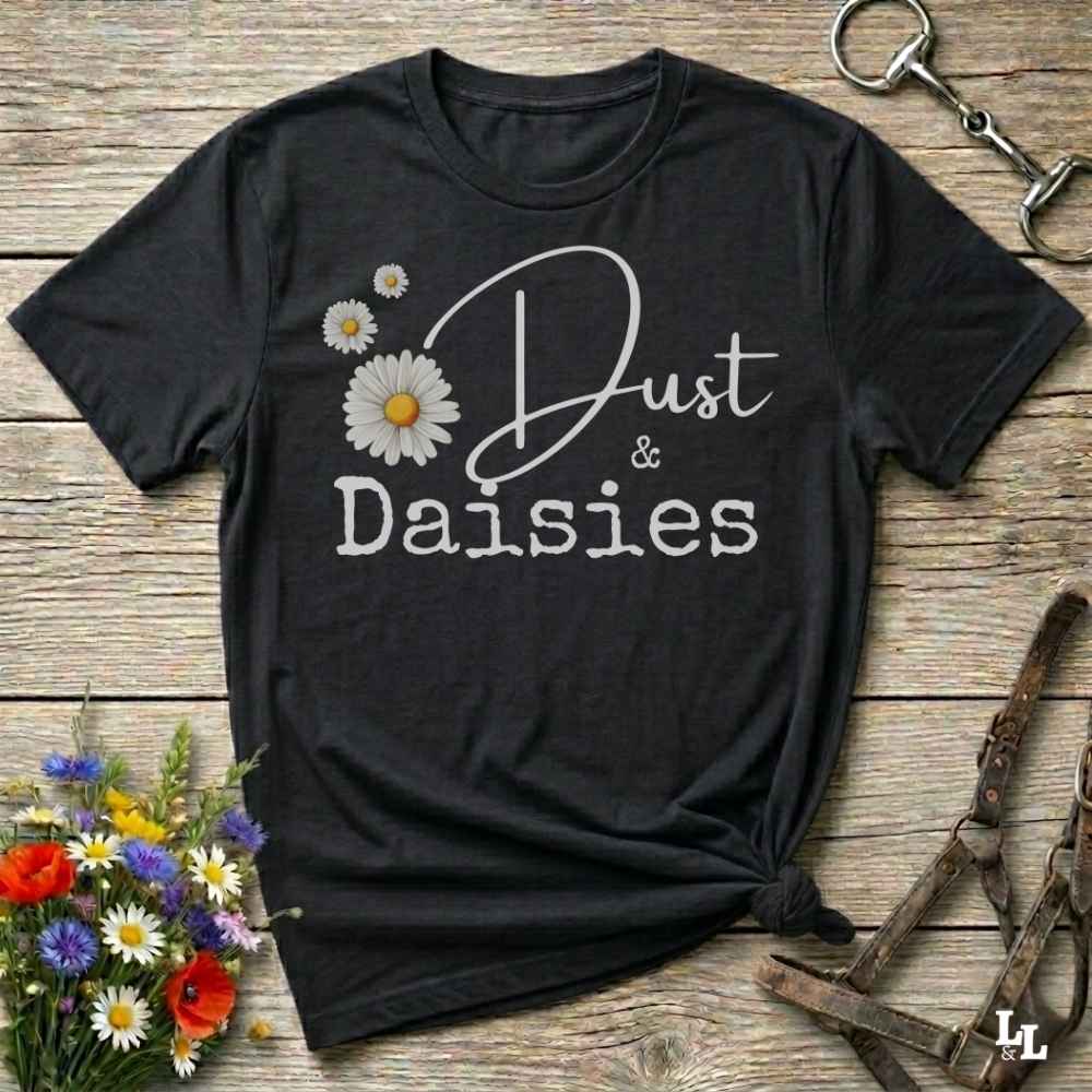A black short-sleeve graphic T-shirt laid flat on a rustic wooden surface, with white script and block text reading 'Dust & Daisies' and white daisy illustrations. The shirt is styled with flowers and equestrian tack props around it.