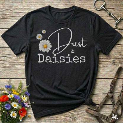 A black short-sleeve graphic T-shirt laid flat on a rustic wooden surface, with white script and block text reading 'Dust & Daisies' and white daisy illustrations. The shirt is styled with flowers and equestrian tack props around it.