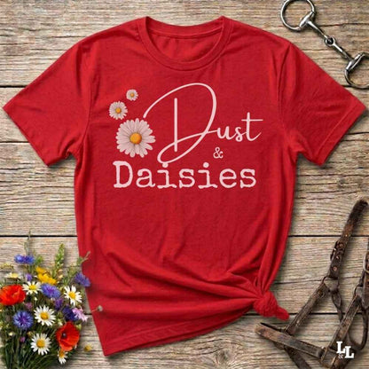 A red short-sleeve t-shirt with a white graphic reading 'Dust & Daisies' and daisy illustrations, styled flat on a rustic wooden surface with flowers and horse tack props.