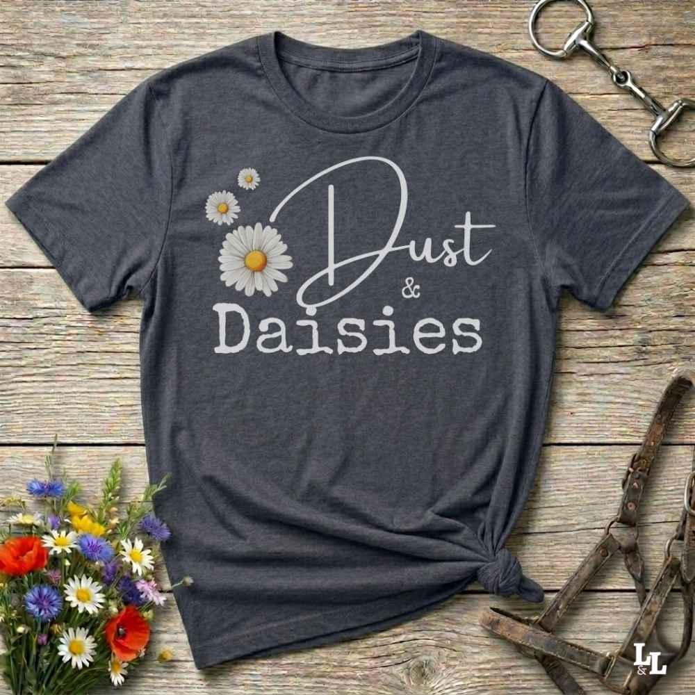A dark heather gray graphic t-shirt laid flat on a rustic wooden surface, styled with equestrian props like a horse bit and halter, and small wildflowers. The shirt features a white graphic print that reads 'Dust & Daisies' accompanied by white and yellow daisy illustrations.