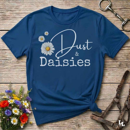 A navy blue short-sleeve t-shirt with a white graphic reading 'Dust & Daisies' and daisy illustrations, styled flat on a rustic wooden surface with flowers and horse tack props.