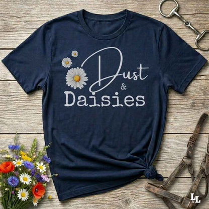 A navy blue short-sleeve t-shirt with a white graphic reading 'Dust & Daisies' and daisy illustrations, styled flat on a rustic wooden surface with flowers and horse tack props.
