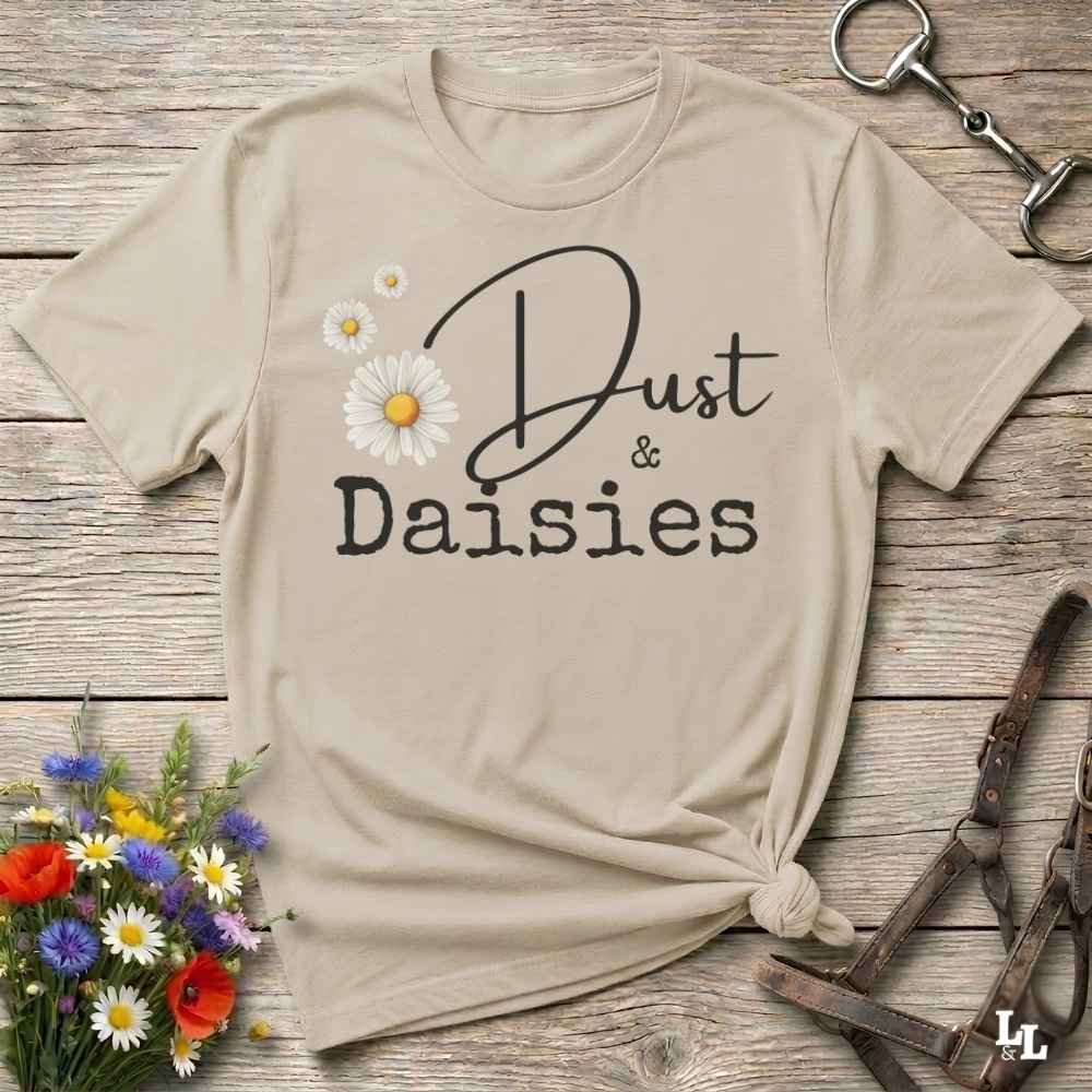 A beige graphic t-shirt with 'Dust & Daisies' printed in black script and block lettering, decorated with white and yellow daisy flowers. The shirt is styled in a flat-lay on a rustic wooden background with a small flower bouquet, a horse bit, and leather tack.