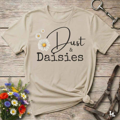 A beige graphic t-shirt with 'Dust & Daisies' printed in black script and block lettering, decorated with white and yellow daisy flowers. The shirt is styled in a flat-lay on a rustic wooden background with a small flower bouquet, a horse bit, and leather tack.