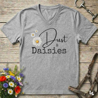 A heather gray V-neck t-shirt with a graphic print reading 'Dust & Daisies' in black script and serif fonts, decorated with white and yellow daisy flowers. The shirt is displayed flat on a rustic wooden background with equestrian-themed props like a horse bit and bridle, and a small bouquet of wildflowers.
