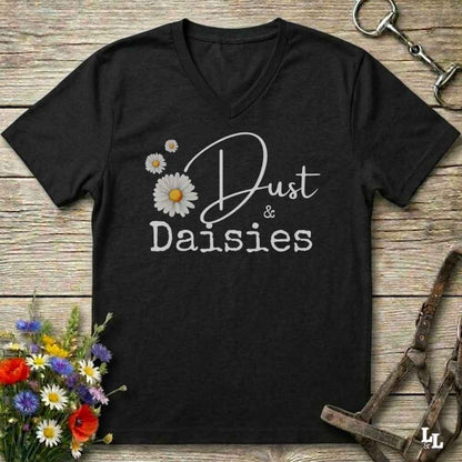 A black V-neck t-shirt with a white graphic reading 'Dust & Daisies' alongside white and yellow daisy illustrations, displayed on a rustic wooden surface with equestrian and floral props.