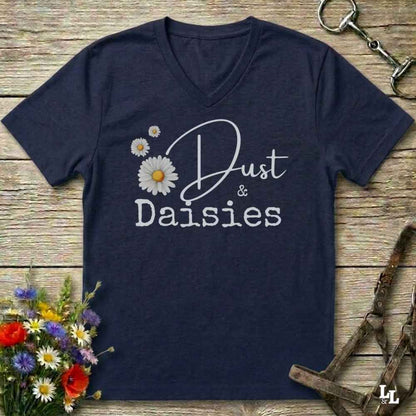 A navy blue V-neck t-shirt with a graphic print reading 'Dust & Daisies' in white script and block lettering, accompanied by three white daisy illustrations. The shirt is displayed flat on a rustic wooden background with equestrian-themed props including a horse bit, leather bridle, and a bouquet of wildflowers.