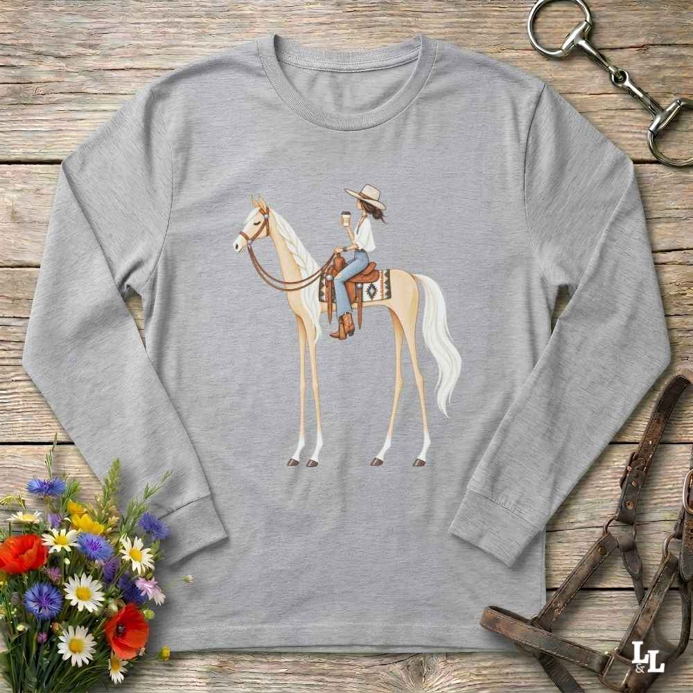 A heather gray long-sleeve t-shirt with a graphic of a cowboy riding a light-colored horse, displayed on a rustic wooden background with decorative floral and equestrian props.