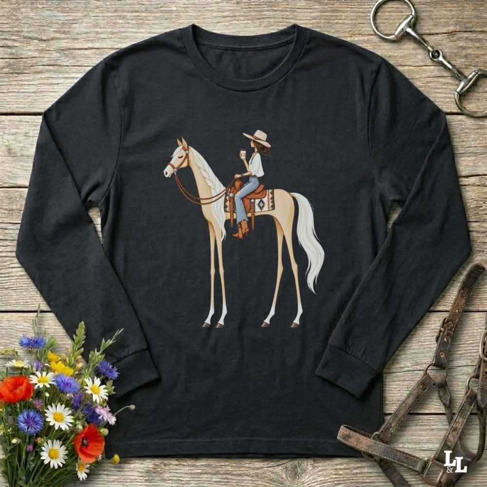 A black long-sleeve crewneck t-shirt featuring a graphic of a cowboy riding a light-colored horse, displayed on a rustic wooden background with equestrian and floral props.