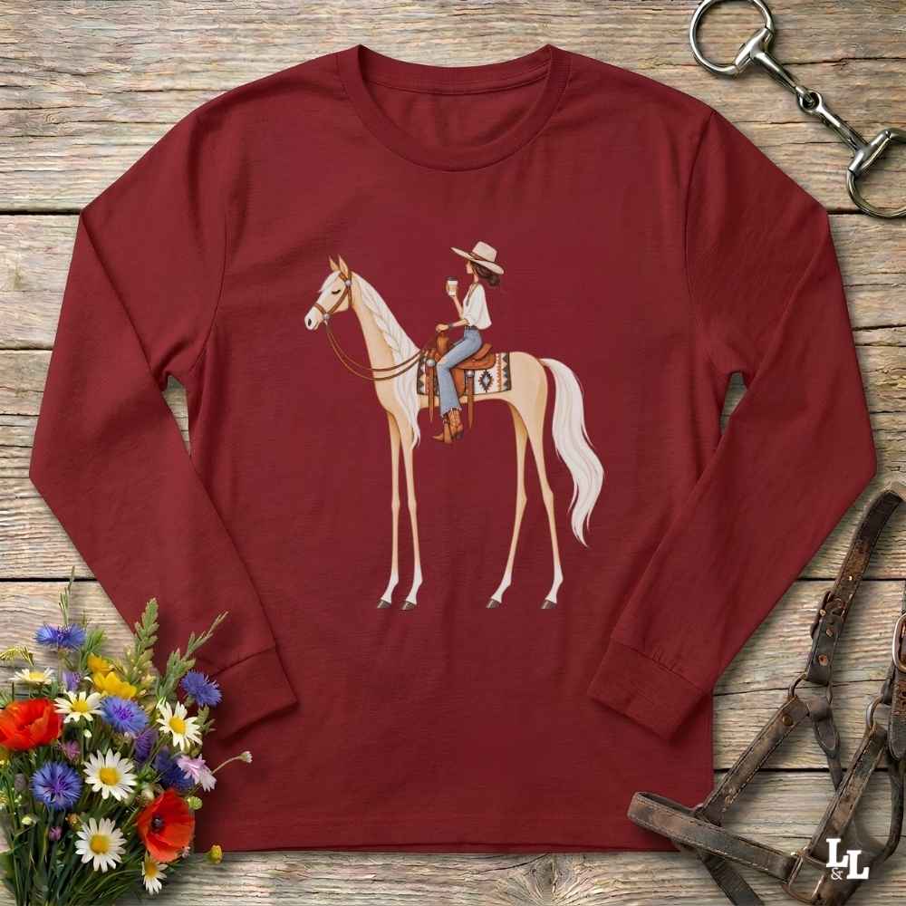A long-sleeve crewneck t-shirt in maroon red, featuring a graphic of a cowboy riding a light-colored horse. The shirt is displayed in a flat-lay arrangement on a rustic wooden background, surrounded by equestrian tack and a small bouquet of wildflowers.