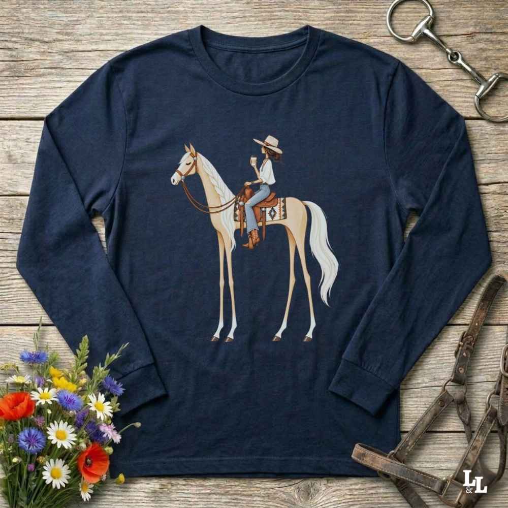 A navy blue long-sleeve t-shirt with a graphic of a cowboy riding a light-colored horse, displayed on a rustic wooden background with equestrian and floral props.