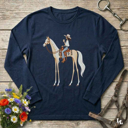 A navy blue long-sleeve t-shirt with a graphic of a cowboy riding a light-colored horse, displayed on a rustic wooden background with equestrian and floral props.