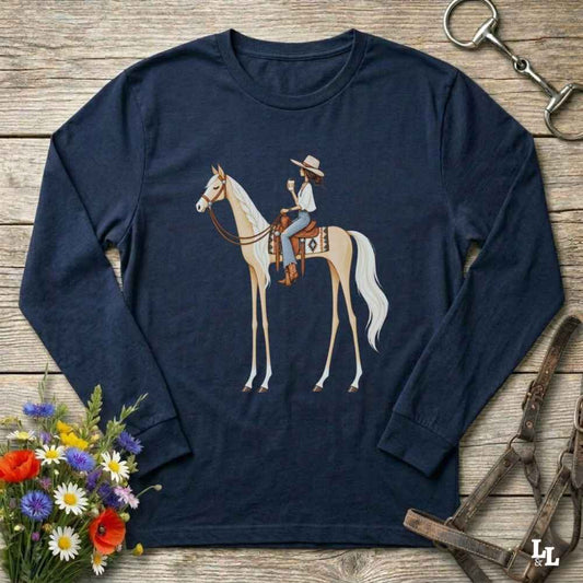 A navy blue long-sleeve t-shirt with a graphic of a cowboy riding a light-colored horse, displayed on a rustic wooden background with equestrian and floral props.