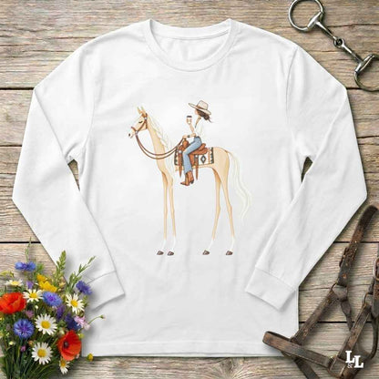 A white long-sleeve crewneck t-shirt with a graphic of a cowboy riding a light tan horse, displayed in a flat-lay arrangement on a rustic wooden background with equestrian accessories and flowers.