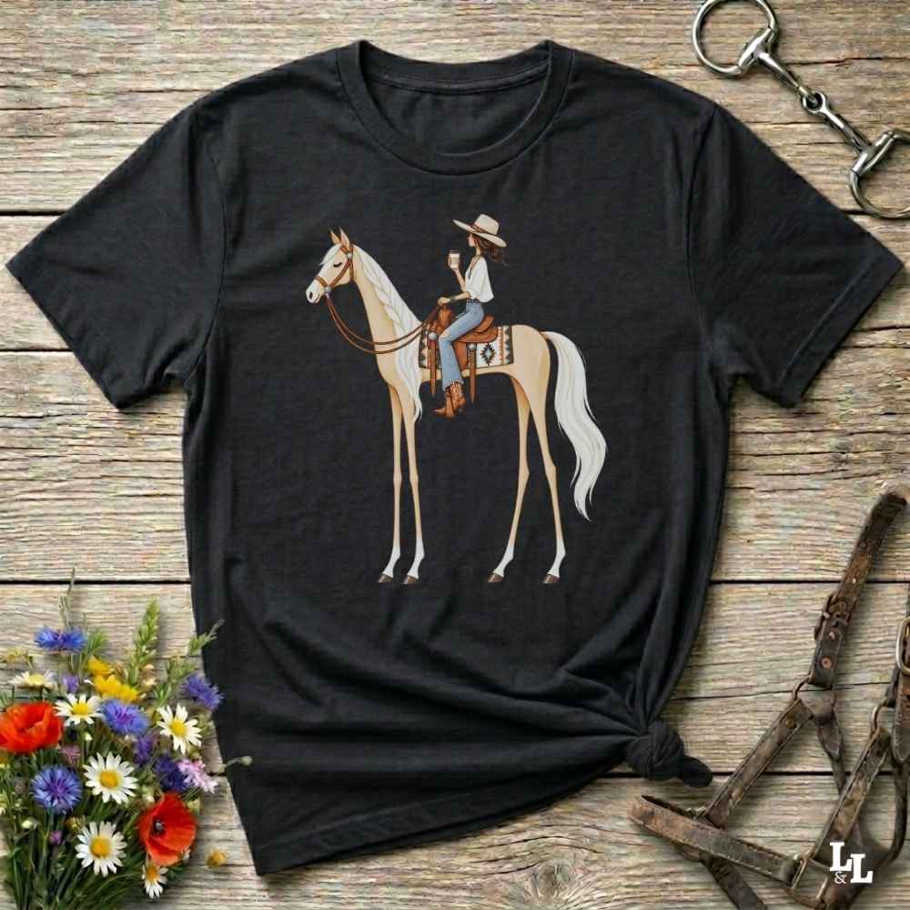 A black short-sleeve graphic T-shirt laid flat on a rustic wooden surface, featuring an illustration of a cowboy riding a light tan horse. The shirt is styled with flowers, a horse bit, and a leather bridle around it.