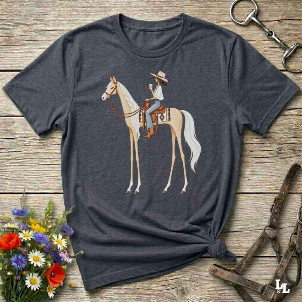 A dark charcoal gray t-shirt with a graphic of a cowboy riding a light tan horse, styled in a flat-lay on a rustic wooden background with equestrian accessories and flowers.
